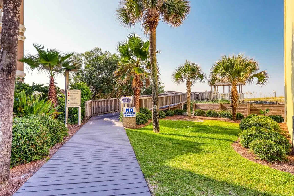 A sun-dappled wooden boardwalk winds through lush palm trees and vibrant tropical landscaping, leading toward a charming gazebo and the promise of a short stroll to the beach. Signs gently remind guests of beach rules, while the inviting path sets a tranquil, vacation-ready mood.