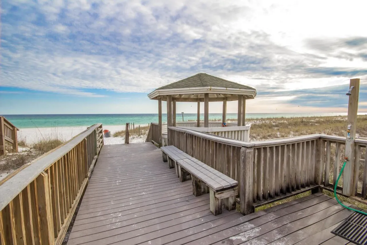 A sunlit wooden boardwalk meanders through lush tropical landscaping, leading directly to a covered beach gazebo. Beyond, white sand stretches to meet the rolling waves of the Gulf, with the ocean horizon line visible. The scene is inviting and promises effortless access to the private beach.