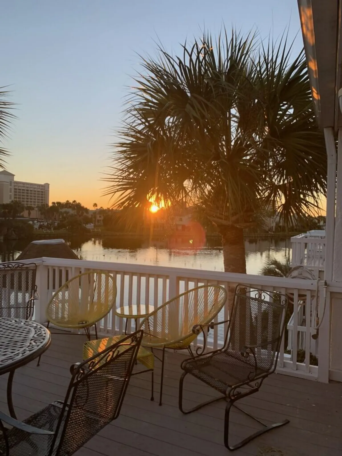 This exterior image captures a stunning sunset over the lake, with a palm tree near the balcony, yellow woven chairs, and metal outdoor furniture set up for relaxation. The scene is peaceful, inviting, and perfectly sets the mood for lakeside serenity and romantic evenings.