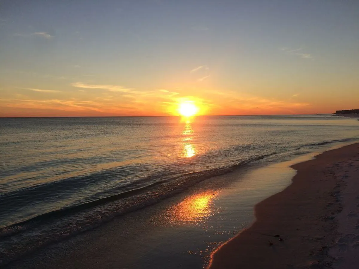 A breathtaking sunset view over a calm ocean, with golden reflections on the water and an empty stretch of sandy coastline. Gentle waves lap at the shore, evoking tranquility and the allure of evening beach walks.