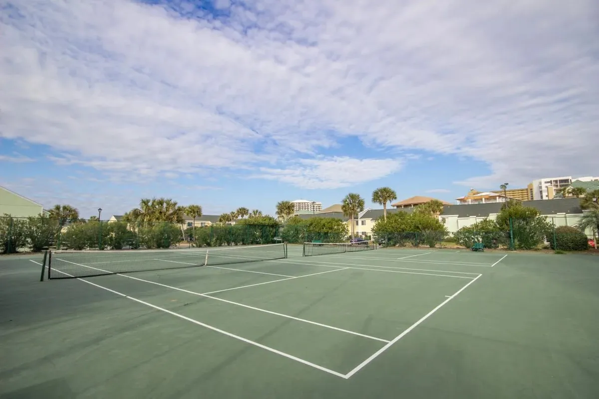 A vibrant exterior shot of the community tennis courts, surrounded by lush green hedges and palm trees, with residential and resort buildings in the background. The courts are well-maintained, with clear court lines and nets, and the setting is bathed in sunlight under a blue sky with scattered clouds. People are visible engaging in tennis play and casual socializing.