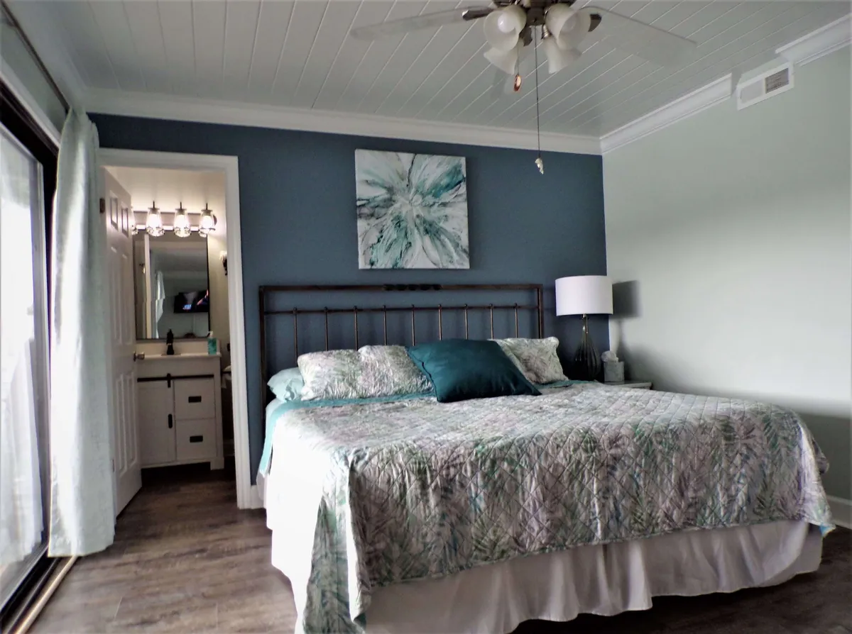 The interior of a tranquil master suite features a king-sized bed with patterned bedding, a deep teal accent wall, and a modern bedside lamp. The bathroom doorway is visible, and there is space ideal for a coffee/tea bar and mini fridge.