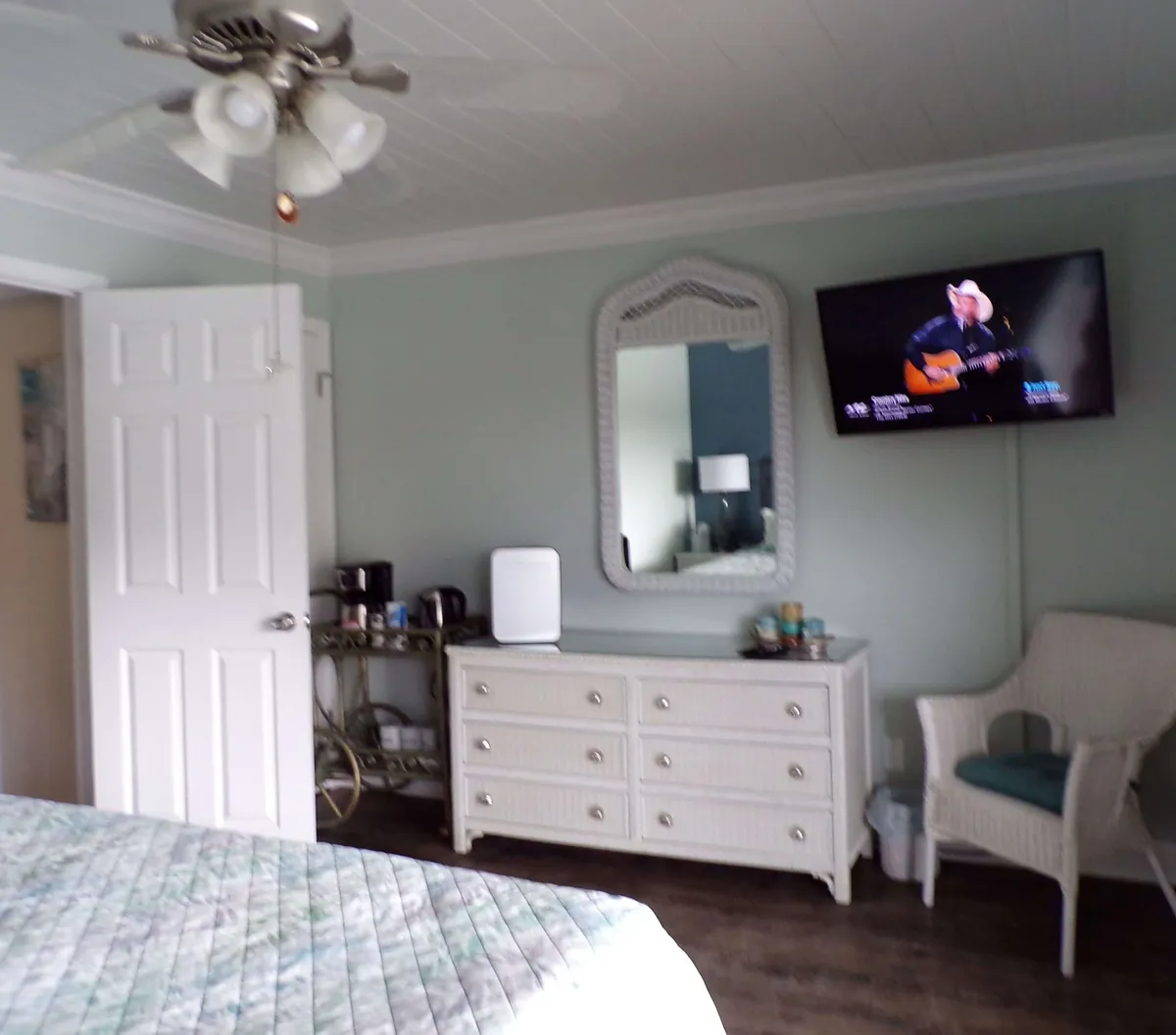 A calming bedroom space featuring a bed with a quilted bedspread, a white wicker dresser with decorative mirror, a wall-mounted flat screen TV, and a cozy wicker chair with cushion. The arrangement suggests modern amenities and a restful retreat, with a hint of a coffee/tea bar area in the room.