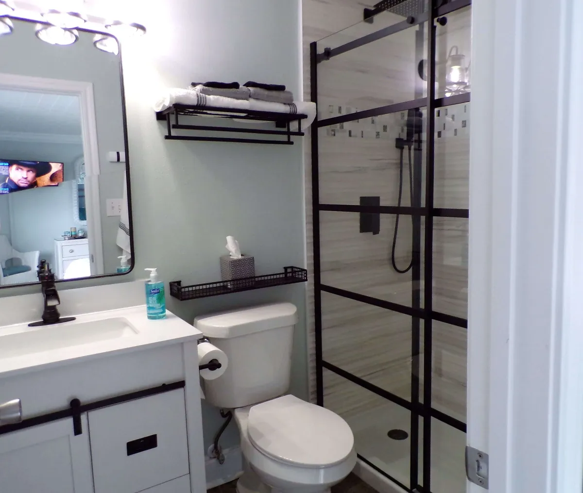A fresh, contemporary bathroom with a modern shower featuring a black grid glass door, white vanity with black faucet and mirror, toilet with tissue holder, and shelves neatly stocked with towels. The space feels organized and inviting.