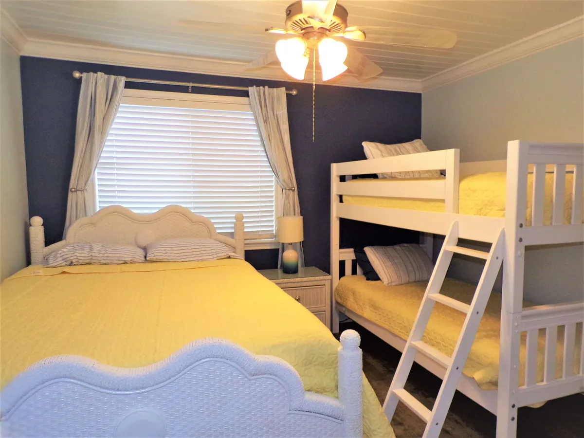 A bedroom featuring a double bed with cheerful yellow bedding, a white bunk bed with a ladder, a window with blinds and curtains, and a ceiling fan with lights. The room exudes a family-friendly, welcoming vibe suited for guests of all ages.