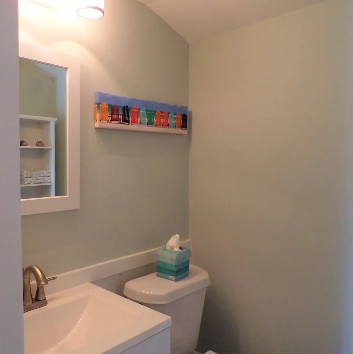 A bright, compact bathroom with a modern white sink and faucet, toilet with a tissue box, colorful beach chair artwork, and a mirror reflecting shelves of towels. The atmosphere is refreshing, clean, and subtly beach-themed.
