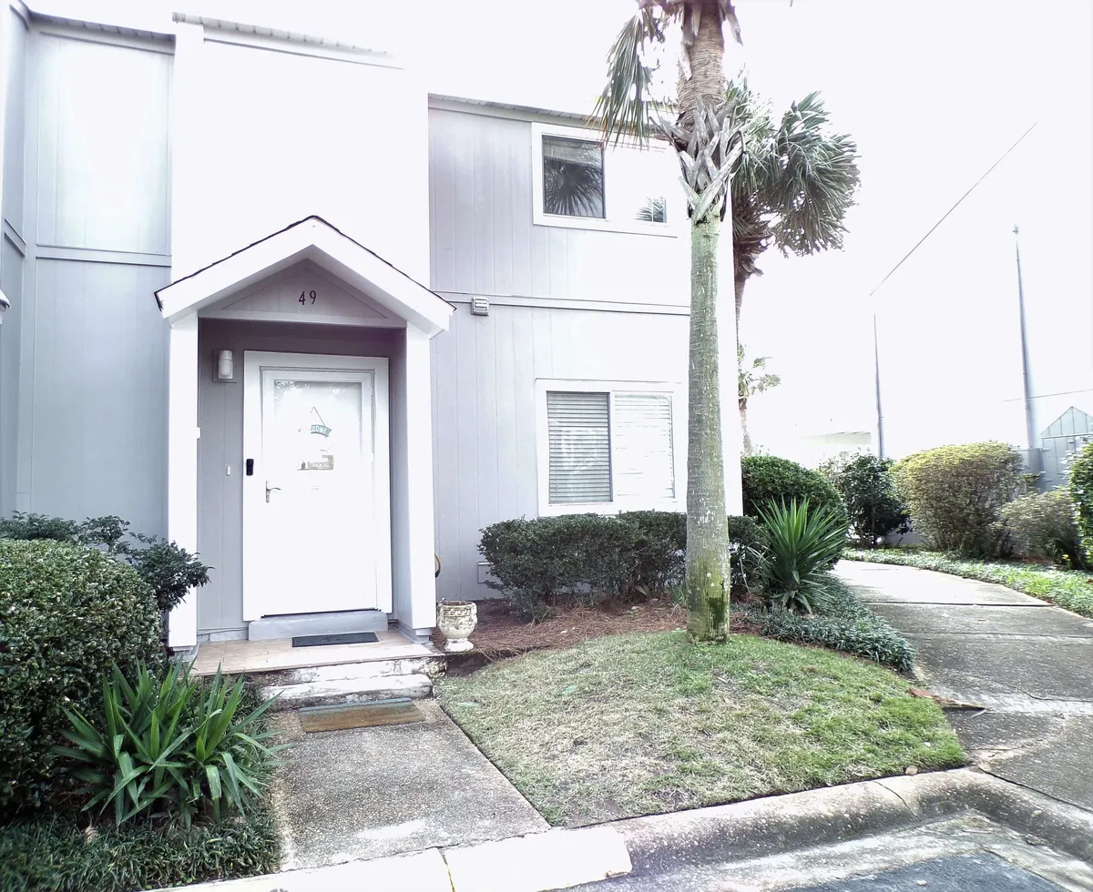 This exterior image features the front entrance with a small porch, palm tree near the walkway, tidy landscaping, and a sidewalk connecting the condo complex. The atmosphere is welcoming and hints at the convenience of beach access and a quiet community.