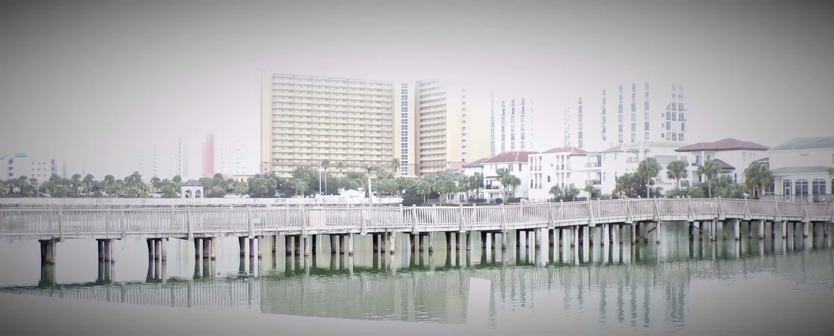 An exterior view of a wooden pier stretching over still water, with high-rise and low-rise buildings in the background. Reflections shimmer on the water’s surface, blending natural and urban scenery. The pier suggests walking, fishing, and access to amenities.