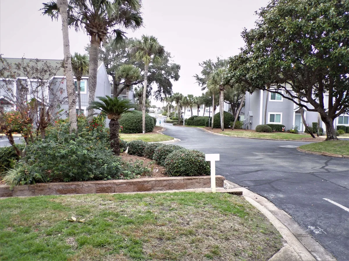 A gently curving road winds through a condo complex bordered by palm trees and manicured gardens. Sidewalks and grassy areas invite leisurely strolls, while the scene suggests easy passage from home to beach with children or gear in tow.