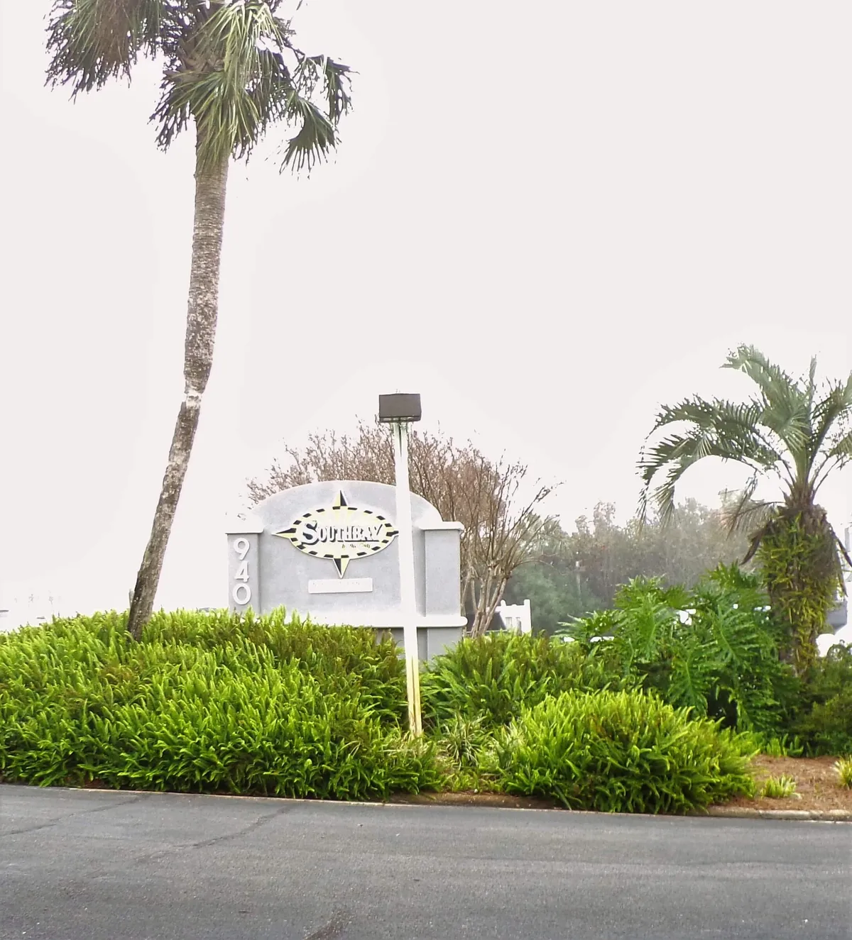 A welcoming entrance sign reading 'Southbay' is nestled among vibrant palm trees and lush tropical shrubs. A paved road leads into the well-maintained condo complex, setting a tranquil and inviting mood.