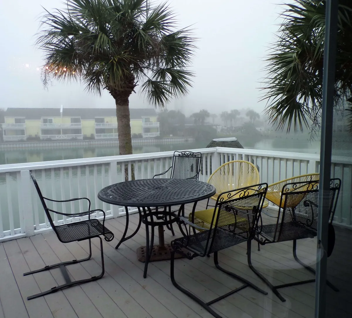 A black metal round table with chairs and yellow accent seating sits on a deck overlooking a calm, misty lake. Palm trees frame the view, and the tranquil water sets a peaceful, contemplative mood.